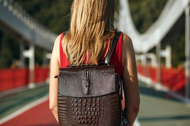 Hübsche Reisende mit einem Lederrucksack, der an einem sonnigen Tag an der Brücke posiert