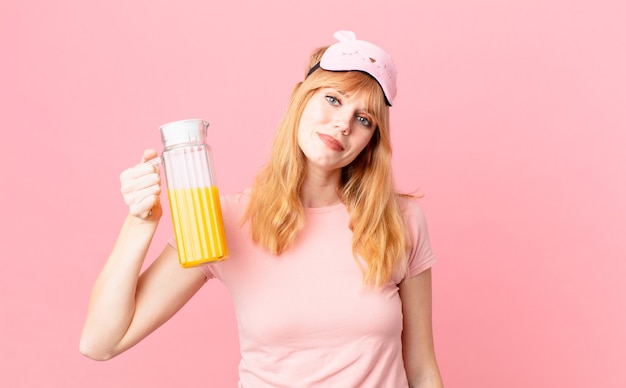 Hübsche rote Kopffrau, die Pyjamas trägt und einen Orangensaft hält. gesundes Frühstückskonzept