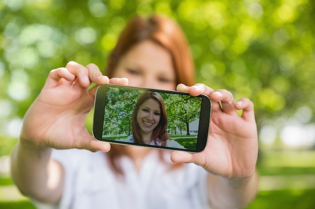 Hübsche Rothaarige, die ein selfie an ihrem Telefon im Park nimmt