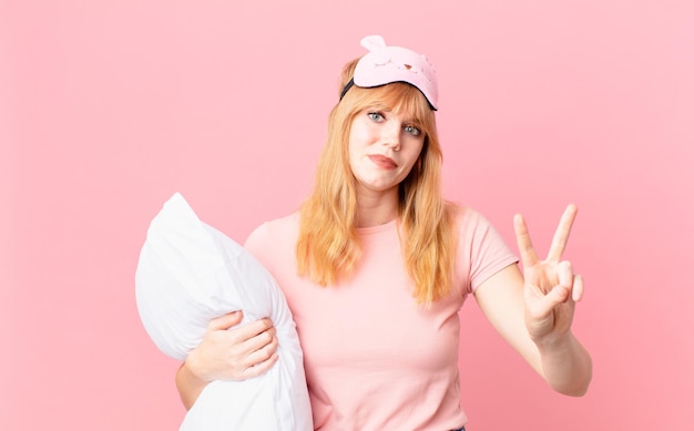 Hübsche rothaarige Frau, die freundlich lächelt und aussieht und Nummer zwei zeigt. Pyjama tragen und ein Kissen halten