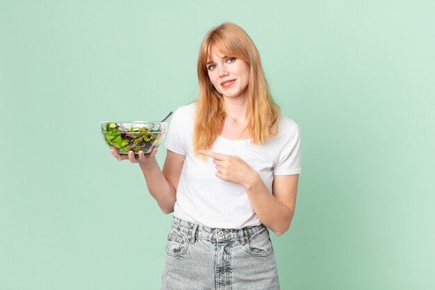 Hübsche rothaarige Frau, die fröhlich lächelt, sich glücklich fühlt und auf die Seite zeigt und einen Salat hält. Diätkonzept