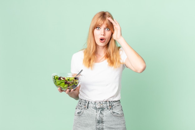 Hübsche rothaarige Frau, die glücklich, erstaunt und überrascht aussieht und einen Salat hält. Diätkonzept