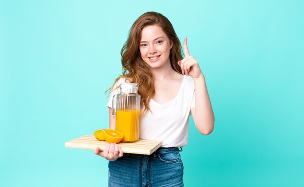 Hübsche rothaarige Frau, die stolz und selbstbewusst lächelt, die Nummer eins macht und einen Orangensaft hält