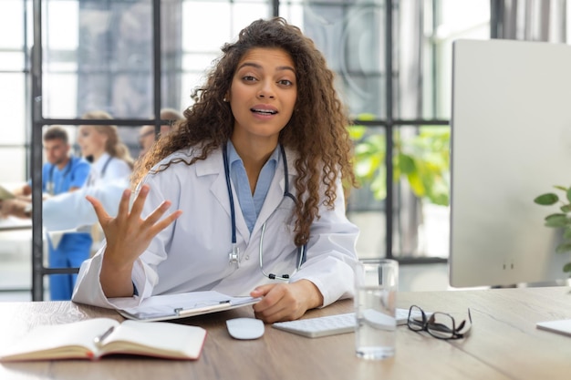 Hübsche Ärztin, die mit einer Webkamera spricht und virtuelle Patienten online per Videokonferenz-Chat berät Kollegen stehen im Hintergrund