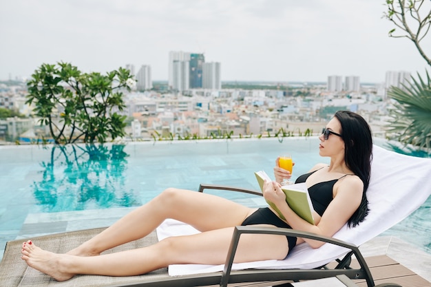 Hübsche schlanke junge Frau, die auf Chaiselongue nahe Infinity-Pool mit Buch und Glas Orangensaft und Blick auf Großstadt entspannt