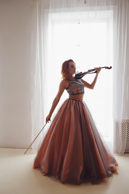 Hübsche schlanke junge Frau mit Geige am Fenster