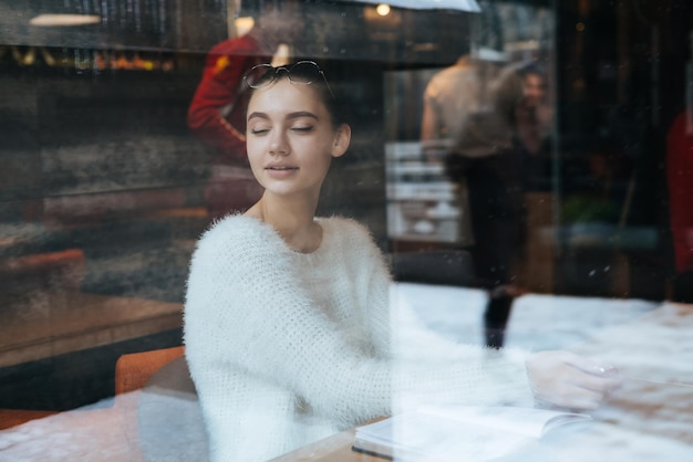 Hübsche schöne Mädchenfreiberuflerin in einer weißen Jacke ruht sich von der Arbeit aus, sitzt in einem gemütlichen Café