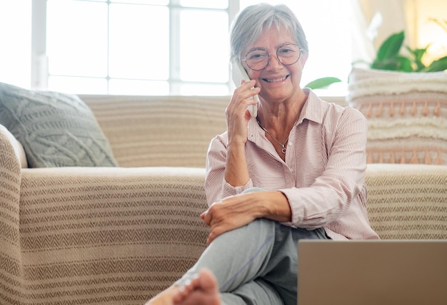 Hübsche Seniorin entspannt auf dem Boden sitzend bei der Heimarbeit und spricht auf dem Smartphone Lächelnde reife Dame, die elektronische Geräte von zu Hause aus verwendet