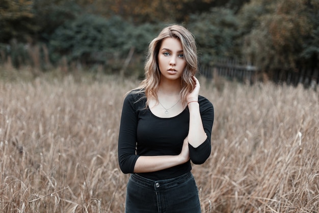 Hübsche sexy junge Frau im schwarzen Vintage-Hemd in den modischen Jeans, die in einem Feld unter trockenem Gras des Herbstes aufwerfen