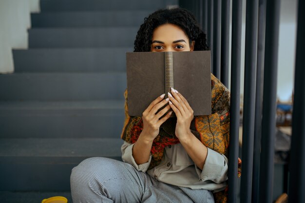 Hübsche Studentin mit Buch, das auf den Stufen im Bibliothekscafé sitzt