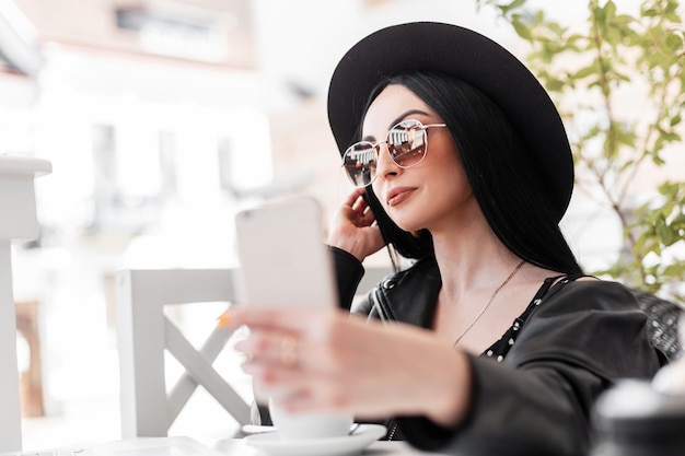 Hübsche stylische Frau mit trendigem Hut in Vintage-Sonnenbrille in modischer Kleidung sitzt im Café und macht Selfie auf dem Smartphone. Attraktives Mädchen in schwarzer Jugendkleidung trinkt Kaffee und fotografiert sich.