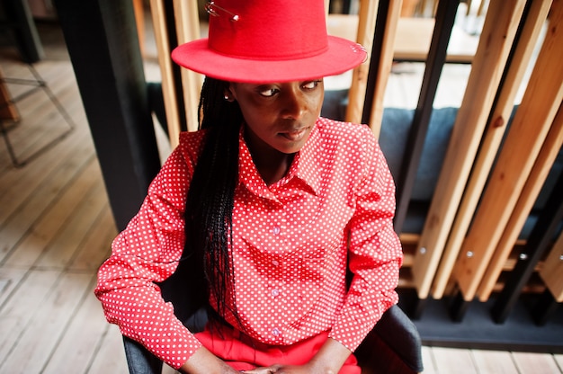 Hübsche Zöpfe Business Afroamerikaner Dame helle herrische Person freundlich tragen Büro rotes Hemd, Hut und Hose, sitzen auf dem Stuhl.