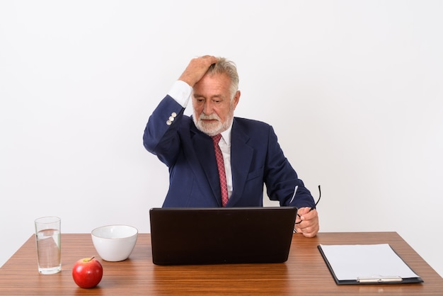 hübscher älterer bärtiger Geschäftsmann, der Brillen hält und Haare zurück bürstet, während Laptop mit grundlegenden Dingen für die Arbeit am Holztisch auf Weiß verwendet.