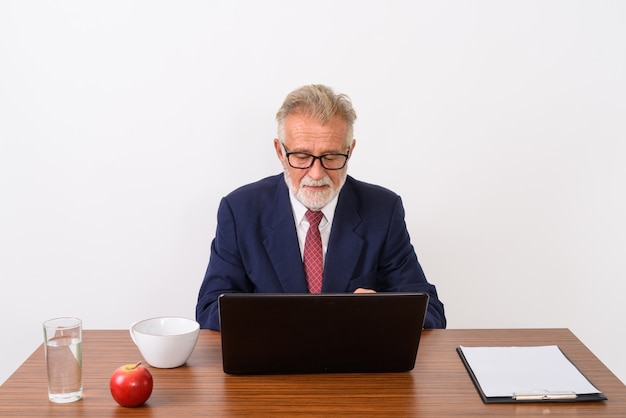 hübscher älterer bärtiger Geschäftsmann, der Laptop verwendet, während er mit grundlegenden Dingen für die Arbeit am Holztisch auf Weiß sitzt