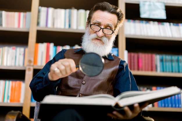 Hübscher älterer bärtiger mann, bibliothekar oder professor, in der bibliothek, sitzend auf dem hintergrund von bücherschränken, hält lupe und lesebuch