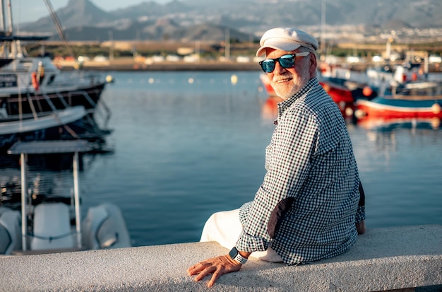 Hübscher älterer Mann in lässigem Hemd und Hut, der bei Sonnenuntergang am Seehafen sitzt