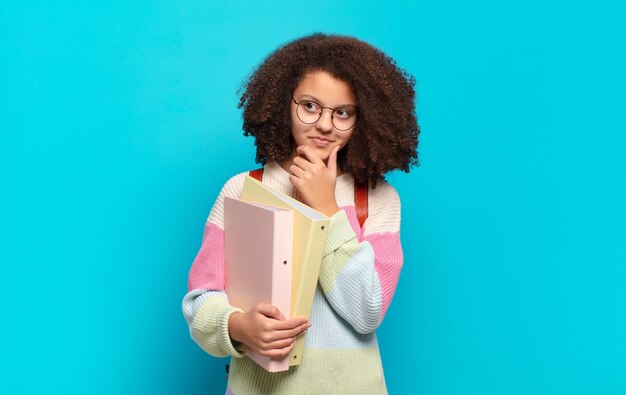 Hübscher Afro-Teenager, der mit einem glücklichen, selbstbewussten Ausdruck mit der Hand am Kinn lächelt, sich wundert und zur Seite schaut. Studentenkonzept