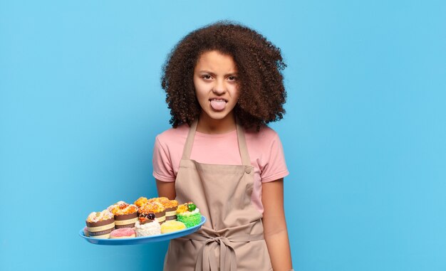 Hübscher Afro-Teenager, der sich angewidert und irritiert fühlt, die Zunge herausstreckt und etwas Böses und Ekelhaftes nicht mag. humorvolles Bäckerkonzept