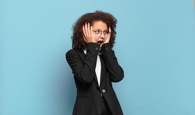 Hübscher Afro-Teenager, der sich glücklich, aufgeregt und überrascht fühlt und mit beiden Händen im Gesicht zur Seite schaut. humorvolles Geschäftskonzept