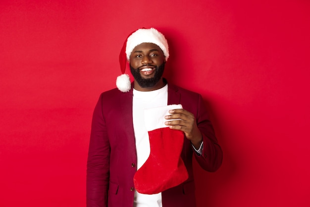 Hübscher Afroamerikaner, der Winterferien feiert, Weihnachtssocke hält und lächelt, Weihnachtsmütze trägt und über rotem Hintergrund steht.