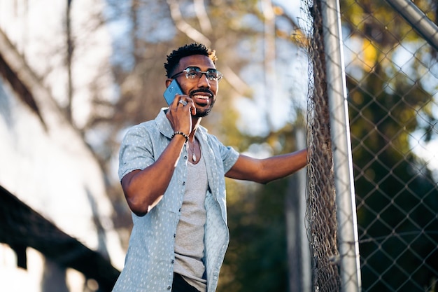 Hübscher afroamerikanischer Mann, der sein Smartphone auf der Straße benutzt und lächelt, lehnt sich an einen Zaun