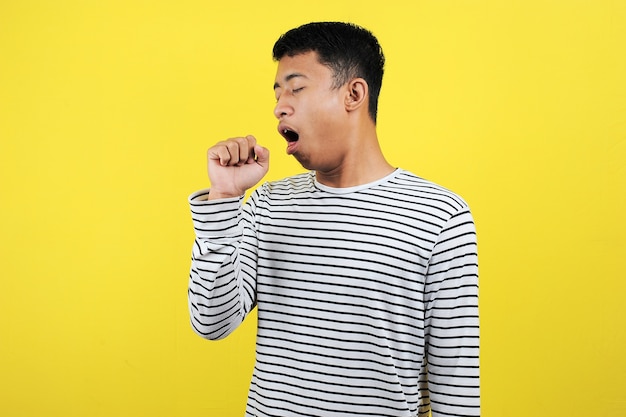 Hübscher asiatischer junger Mann, der sich unwohl fühlt und als Symptom für Erkältung oder Bronchitis hustet. Gesundheitskonzept isoliert auf gelbem Hintergrund