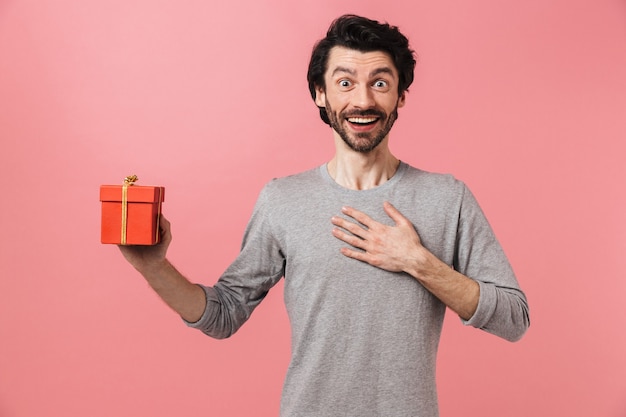 Hübscher aufgeregter junger bärtiger brünetter Mann, der Pullover trägt, der über rosa steht und Geschenkbox hält