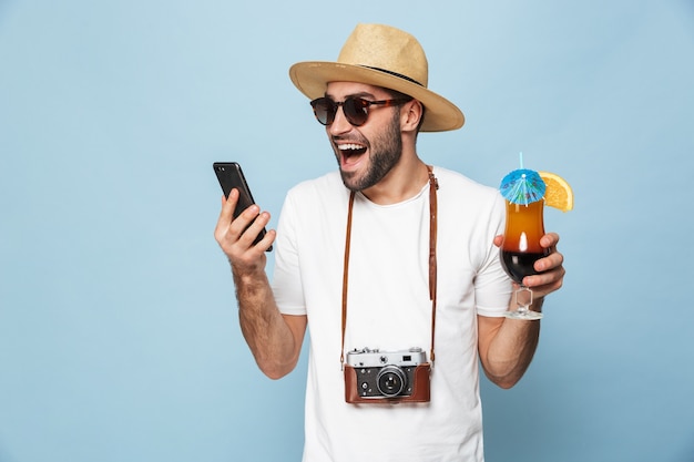 hübscher aufgeregter junger Mann Tourist posiert mit Kamera mit Handy trinken Cocktail über blaue Wand isoliert.