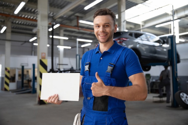 Hübscher Automechaniker mit dem weißen leeren Brett in der Hand posiert gegen den modernen Reparaturautoservice