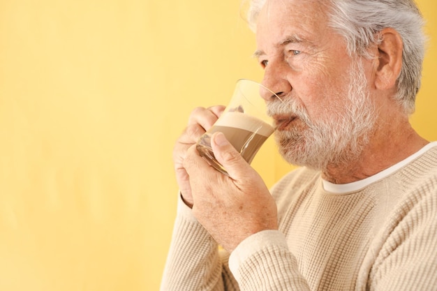 Hübscher bärtiger älterer Mann, der Kaffee und Milchcappuccino aus einem Becherglas trinkt und auf gelbem Hintergrund steht