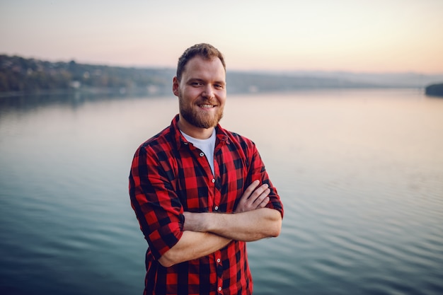 Hübscher bärtiger blonder kaukasischer Mann, der auf Klippe mit vom Fluss gekreuzten Armen steht.