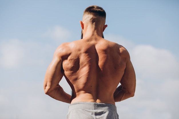 Hübscher bärtiger Bodybuilder-Mann mit perfektem Muskelkörper, der draußen auf dem Himmelhintergrund aufwirft. Starker Mann mit nacktem Oberkörper, der wegschaut. Blick von hinten