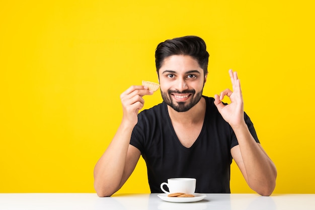 Hübscher bärtiger indischer Mann, der gesunde Vollkornkekse isst, die in Kaffee von Tee oder Chai in einer Tasse getaucht sind und am Tisch vor gelbem Hintergrund sitzen