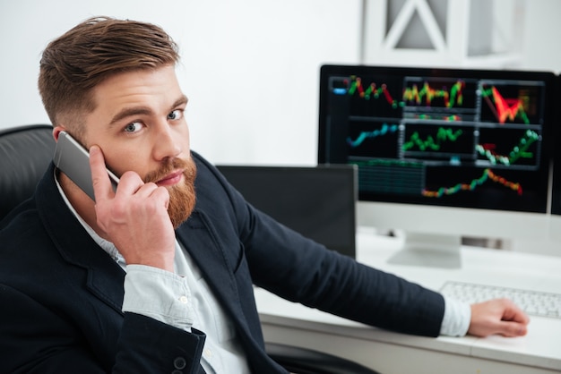 Hübscher bärtiger junger Geschäftsmann, der im Büro am Handy spricht