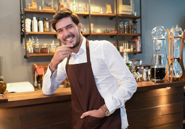 Hübscher bärtiger Mann in Schürze, der in die Kamera blickt und die Hand am Kinn hält, während er an der Bartheke steht Startup-Kleinunternehmer-Konzept