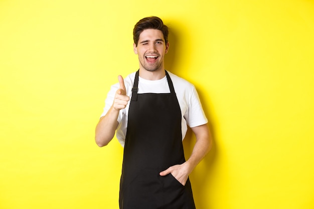 Hübscher Barista in schwarzer Schürze begrüßt die Gäste im Café, zeigt mit der Fingerpistole und zwinkert und steht vor gelbem Hintergrund.