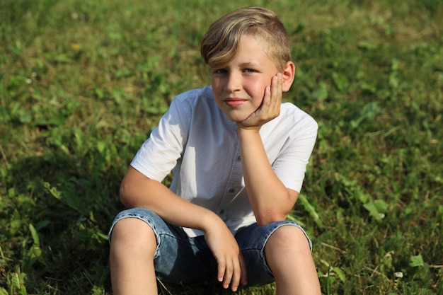 Hübscher blonder Junge, der im Sommer im Park auf dem Gras sitzt. Foto in hoher Qualität