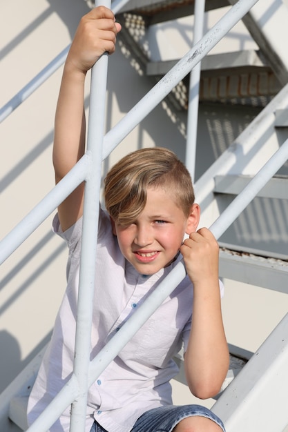 Hübscher blonder Junge in einem weißen Hemd, das im Sommer auf einer weißen Treppe lächelt. Foto in hoher Qualität