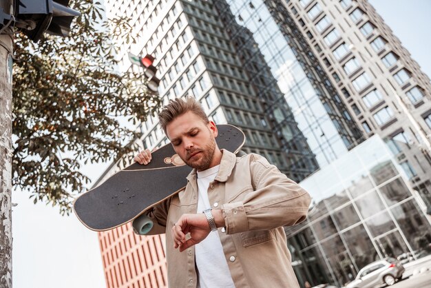 Hübscher blonder junger Mann Skateboarder, der Longboard auf der Schulter in Eile hält und ernst auf die Armbanduhr schaut. Unterwegs städtischer Gebäudehintergrund. Trägt ein beigefarbenes Jeanshemd. Freizeit im Freien. Pendeln