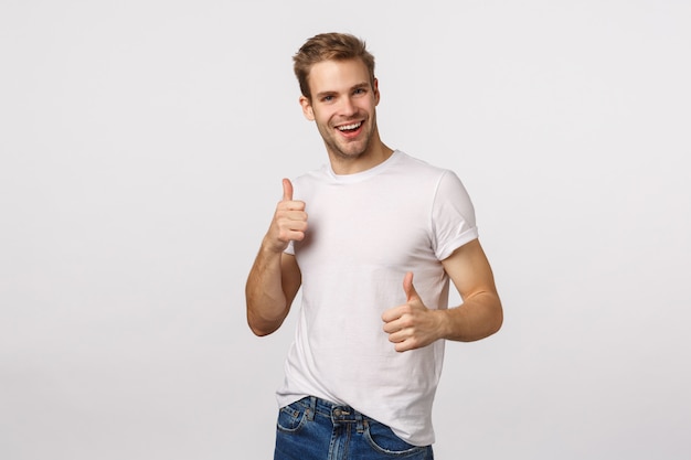 Hübscher blonder Kerl mit blauen Augen und weißem T-Shirt, die Daumen aufgibt