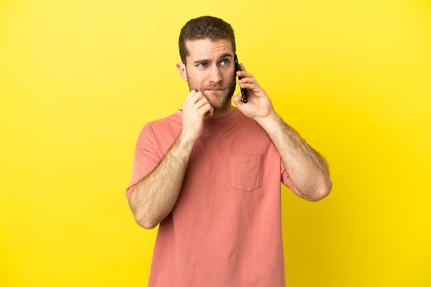 Hübscher blonder Mann, der Handy über isoliertem Hintergrund verwendet, der Zweifel und Denken hat