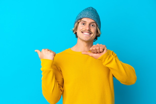 Hübscher blonder Mann lokalisiert auf blauer Wand, die zur Seite zeigt, um ein Produkt zu präsentieren