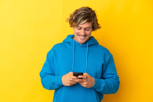 Hübscher blonder Mann lokalisiert auf Gelb, der eine Nachricht mit dem Handy sendet