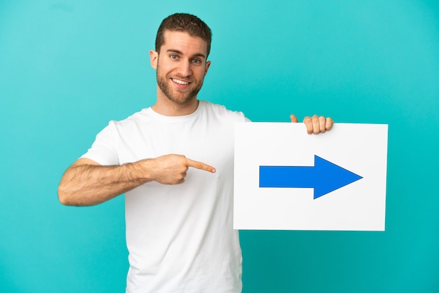 Foto hübscher blonder mann über isoliertem blauem hintergrund, der ein plakat mit pfeilsymbol hält und darauf zeigt