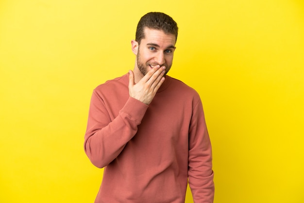 Hübscher blonder Mann über isoliertem gelbem Hintergrund glücklich und lächelnd, den Mund mit der Hand bedeckend