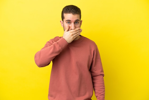 Hübscher blonder Mann über lokalisiertem gelbem Hintergrund, der Mund mit Hand bedeckt