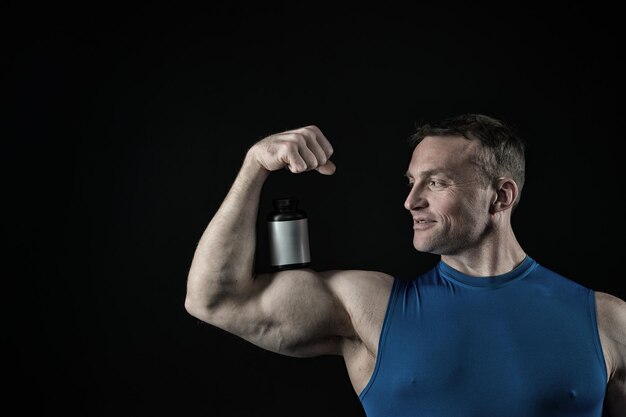 Hübscher Bodybuilder oder sexy muskulöser lächelnder Kerl in blauer Weste mit Sportlerkörper, Torso und Brusttrainingssport im Fitnessstudio, hält Pillendose für Drud, Steroid, Vitamin oder Doping auf schwarzem Hintergrund