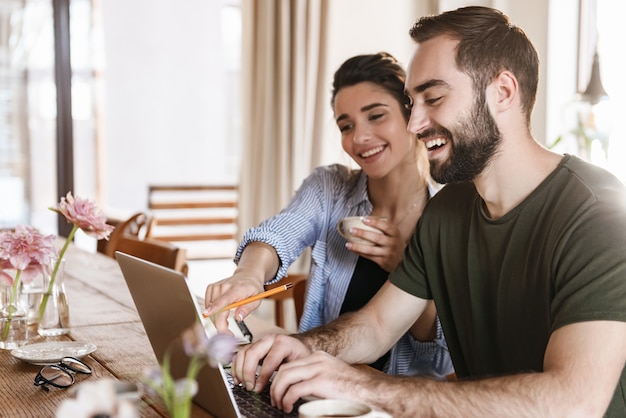 hübscher brünettes Paar Mann und Frau, die Kaffee trinken und zusammen am Laptop arbeiten, während sie zu Hause am Tisch sitzen