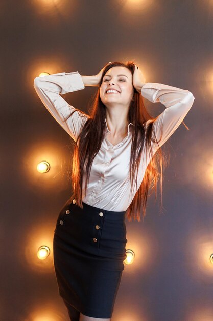 Hübscher Brunette in Bluse und kurzem Rock steht im Studio mit Backsteinmauer und Lampen und hält seine Hände auf seinem Kopf