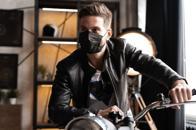 Hübscher brutaler männlicher Biker in der schwarzen Maske in der Lederjacke, die auf dem Motorrad sitzt und nach vorne schaut